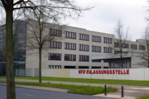 Ein modernes, helles Bürogebäude mit rot gerahmten Fenstern und kahlen Bäumen davor. Ein weißer Zaun trägt das rote Schild "KFZ-ZULASSUNGSSTELLE", das auf eine Kfz-Zulassungsstelle hinweist, bei der die Bearbeitungszeit Zulassungsstelle Berlin beantragt wird.
