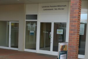 Eingang des Landratsamtes Potsdam-Mittelmark und der Zulassungsstelle Werder in Deutschland, mit Glastüren, Beschilderung darüber und einer Backsteinsäule auf der rechten Seite. Ein freistehendes Schild steht sichtbar davor.